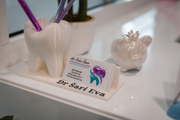 Dental office counter with a stand holding business cards containing the clinic's contact information, a tray with the inscription 'Dr. Eva Sari', a decorative pen holder, and a pig figurine.