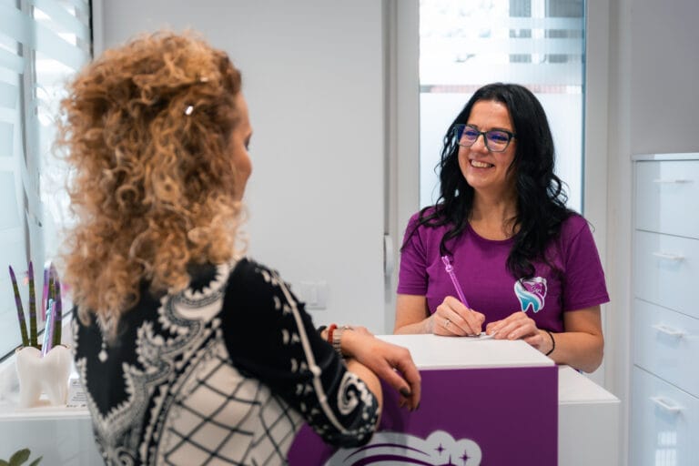 Dr. Eva Sari standing at the dental office counter, writing notes and welcoming a patient.